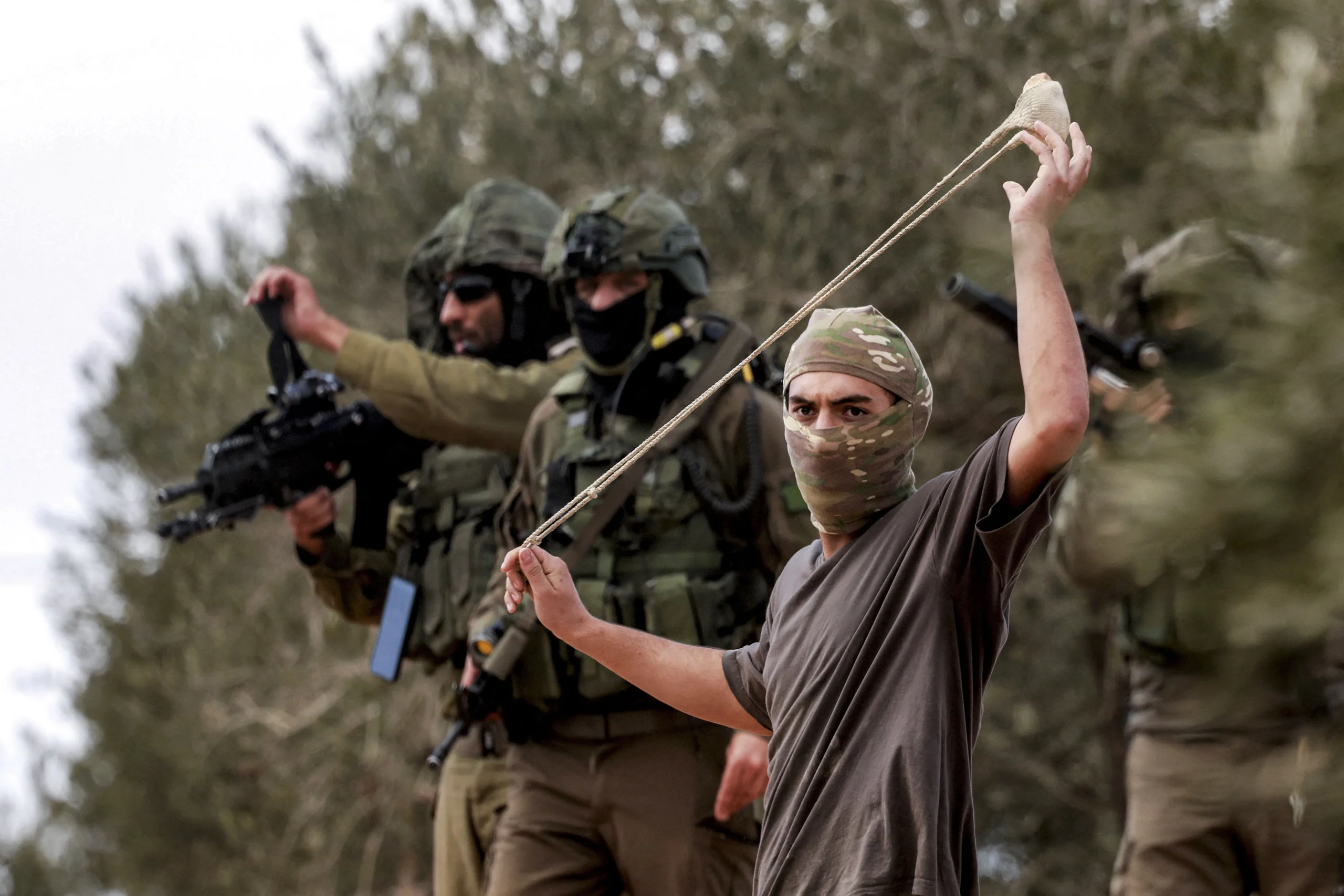 Żołnierze Sił Obronnych Izraela ochraniają zamaskowanego terrorystę osadnika przygotowującego się do wystrzelenia kamienia z procy w kierunku Palestyńczyków zbierających oliwki na południe od Nablusu, na nielegalnie okupowanym Zachodnim Brzegu Palestyny, 10 października 2025 r. (Fot. Jaafar Ashtiyeh / AFP via Getty Images)